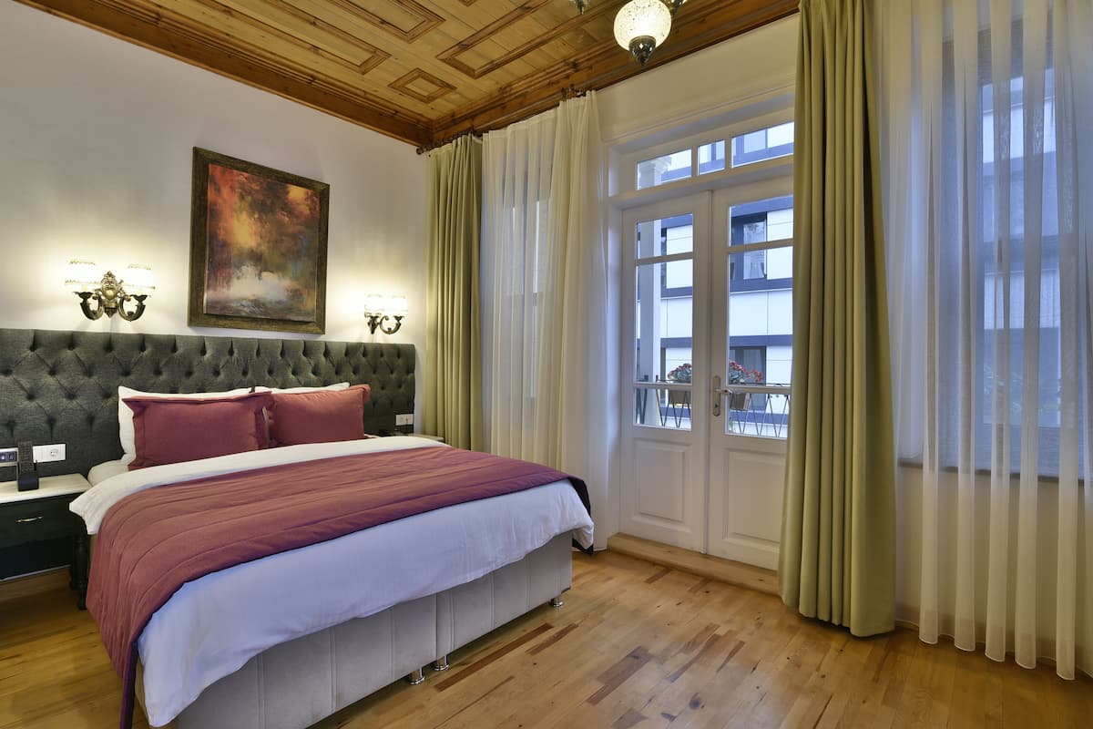 A bedroom with a large bed, a tufted headboard, two bedside lamps, a painting, and a window with curtains.