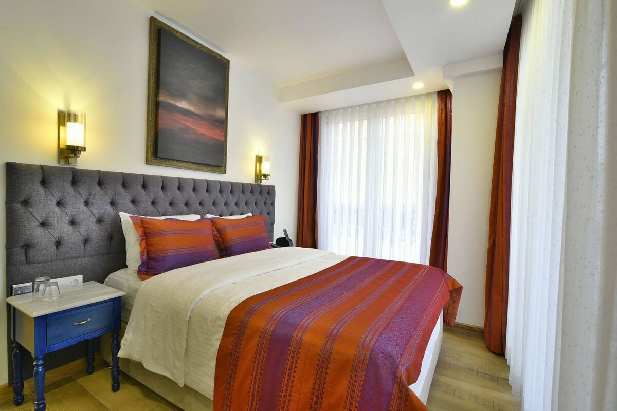 A hotel room with a tufted headboard, a large bed with a patterned bedspread, a bedside table with a lamp, and a painting on the wall.