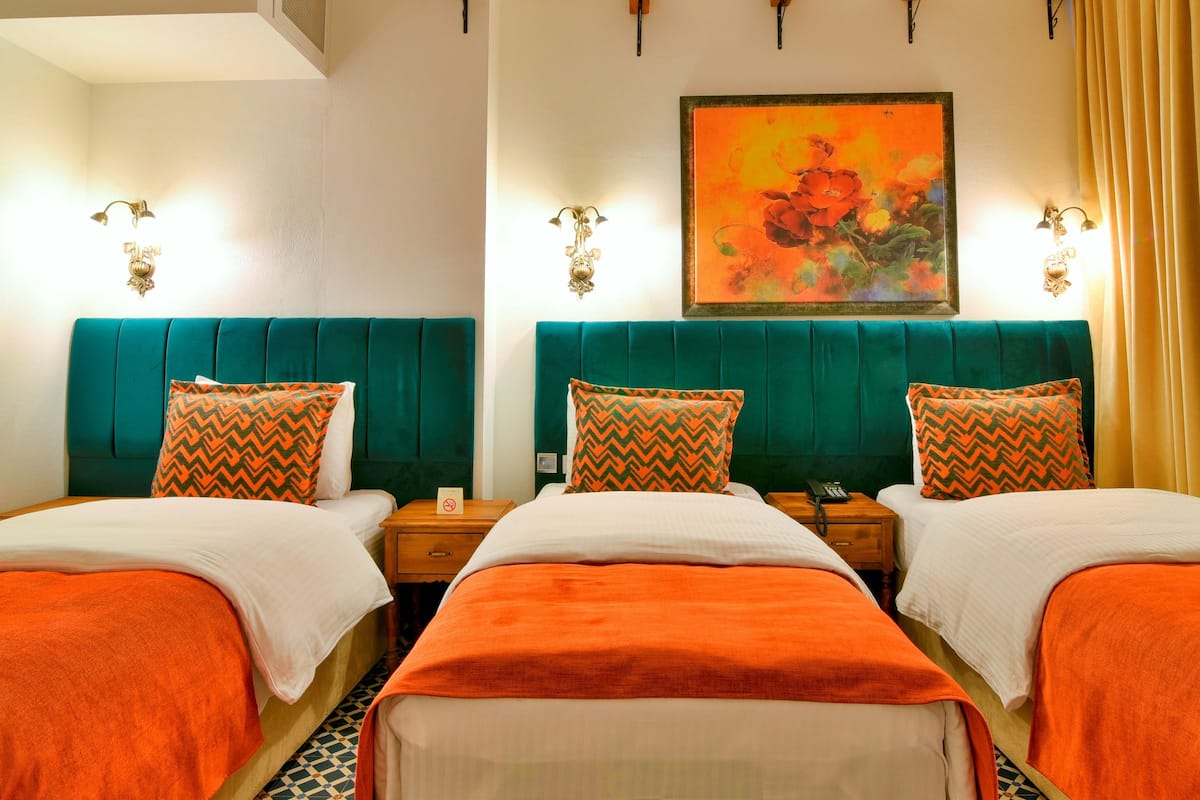 A hotel room with two beds, teal headboards, orange and white bedding, a painting on the wall, and a nightstand with a phone.