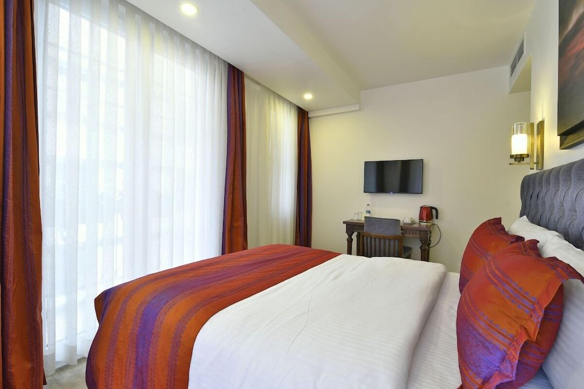 A hotel room with a large bed, a desk with a chair, a flat-screen TV mounted on the wall, and a window with curtains.