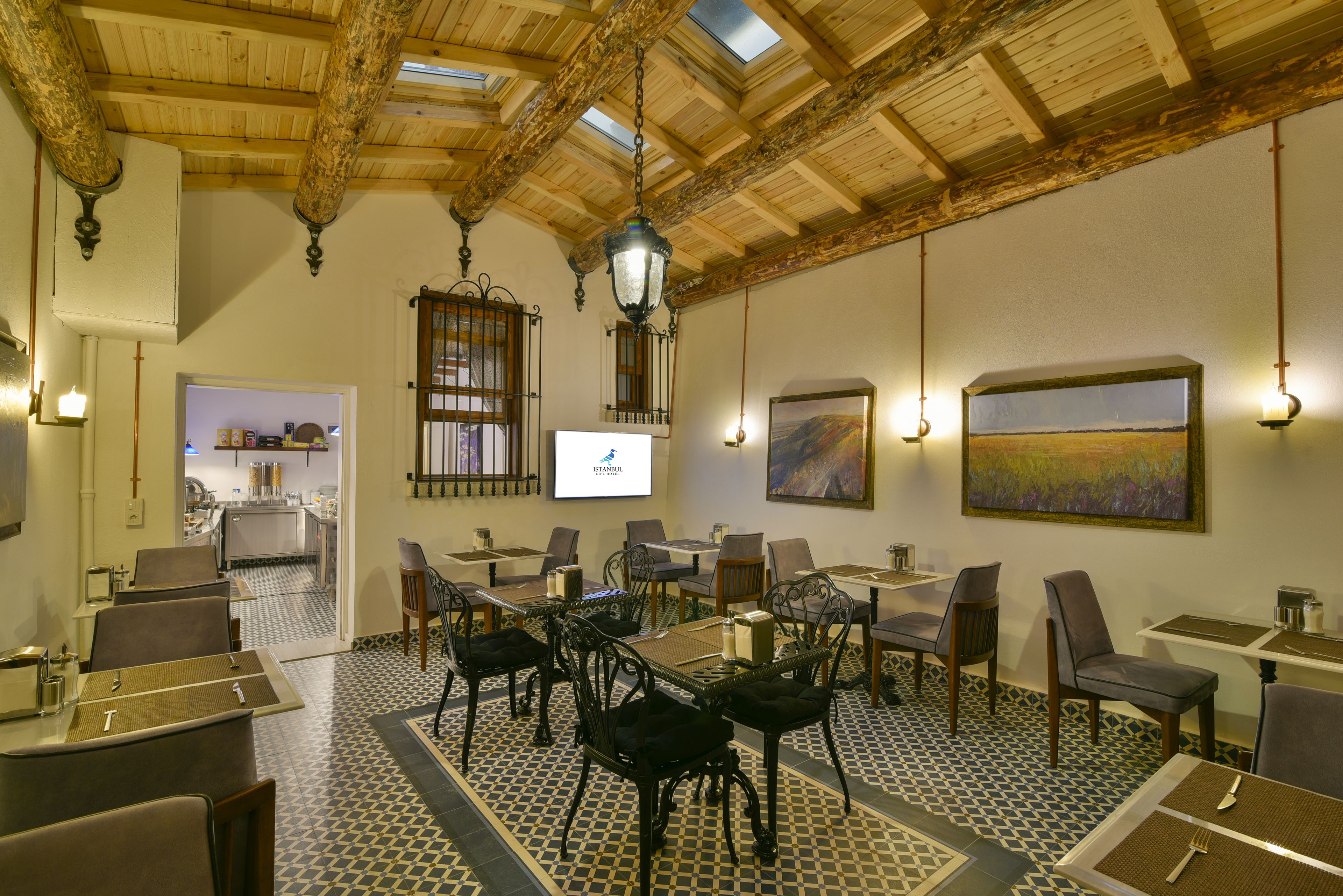 A dining area with tables and chairs, a kitchen in the background, and a painting on the wall.