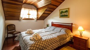 Un dormitorio con techo de madera, una cama con colcha estampada, una mesita de noche con lámpara, una silla y un cuadro en la pared.