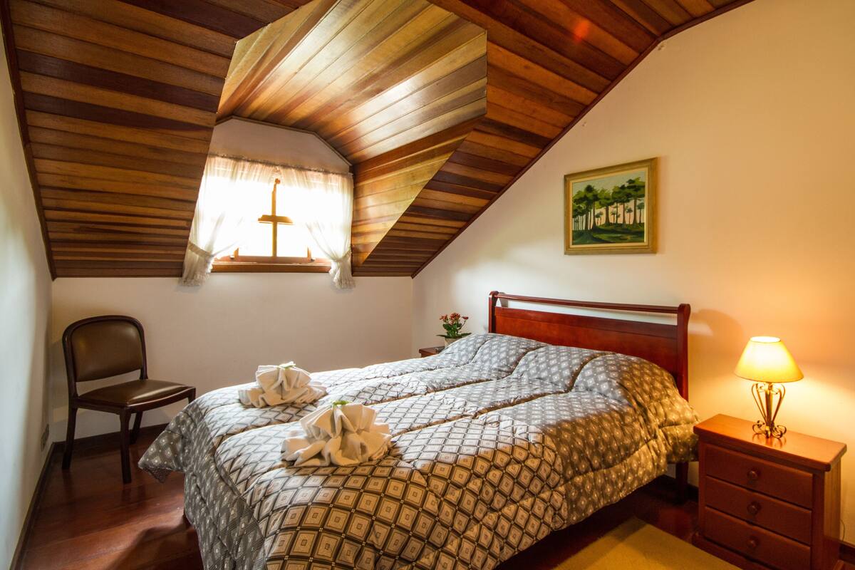 Ein Schlafzimmer mit Holzbalkendecke, einem Bett mit gemustertem  Überwurf, einem Nachttisch mit Lampe, einem Sessel und einem Gemälde an der Wand.