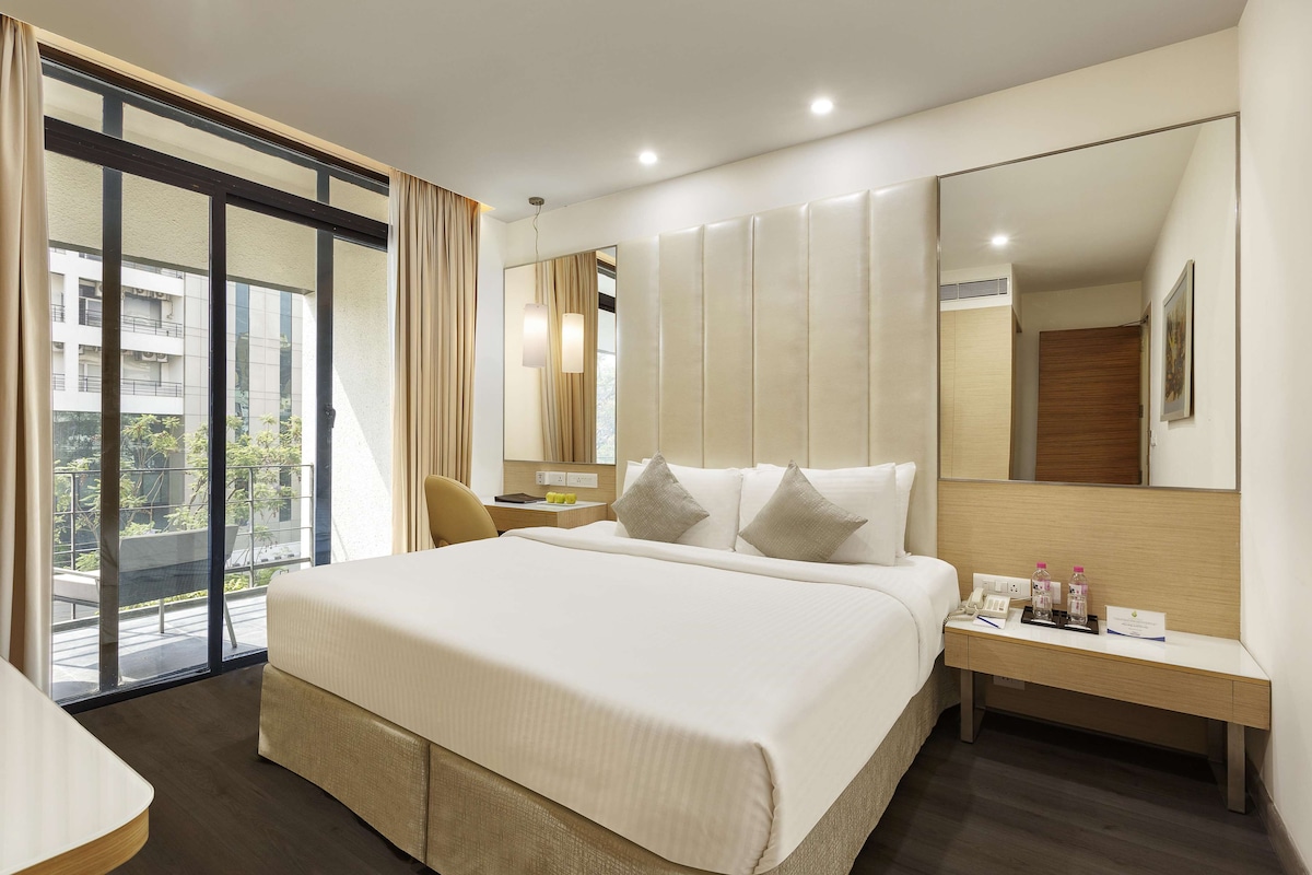 Una habitación de hotel moderna con una cama grande, una mesita de noche con detalles, un espejo y vistas al exterior.