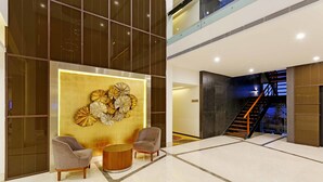 A modern lobby with a large wall art piece, two armchairs, a round wooden table, and a staircase leading to an upper level.