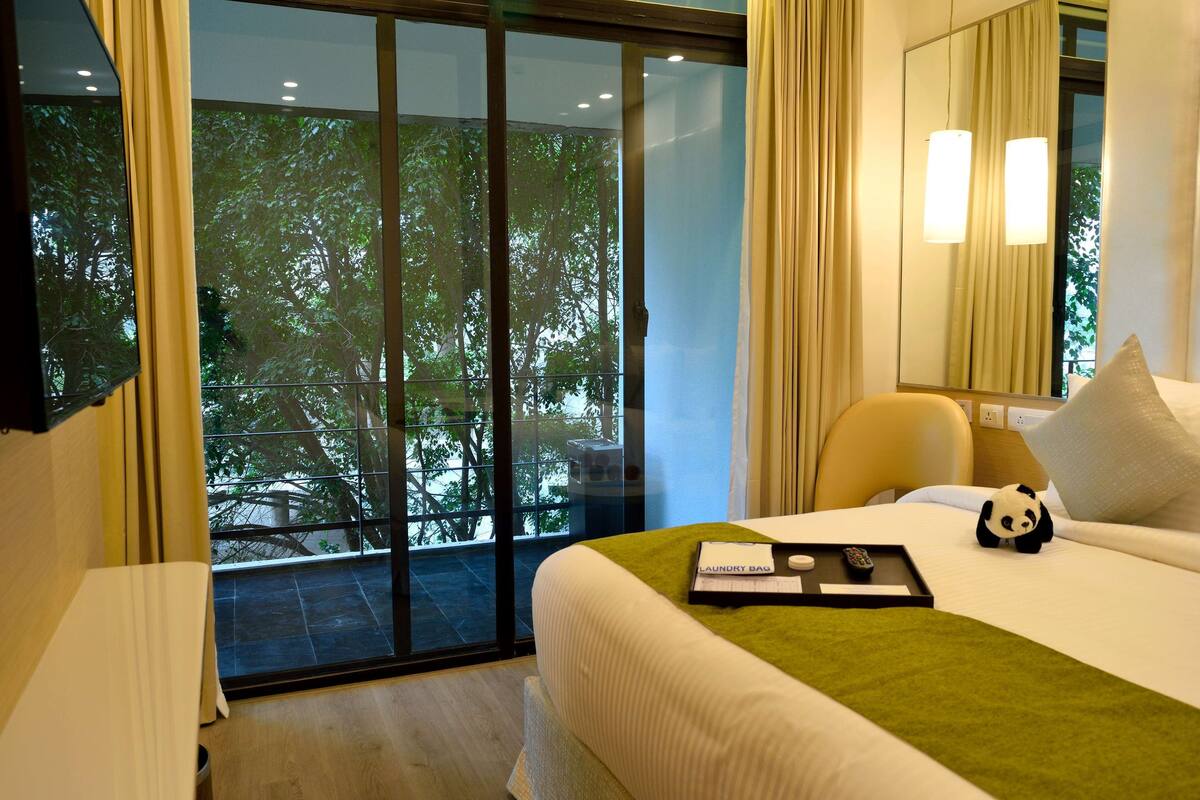 A hotel room with a bed, a chair, a small table with a remote control and a tray, and a large window with a view of trees.