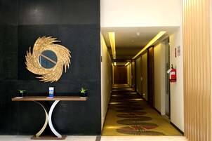 A hotel lobby with a reception desk, a decorative wall piece, a patterned carpet, and a hallway leading to rooms.