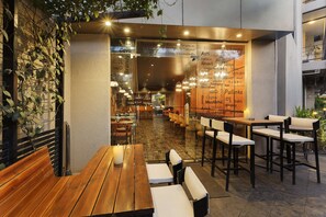 An outdoor dining area with wooden tables and benches, a brick wall with climbing plants, and a menu board with various food and drink options.