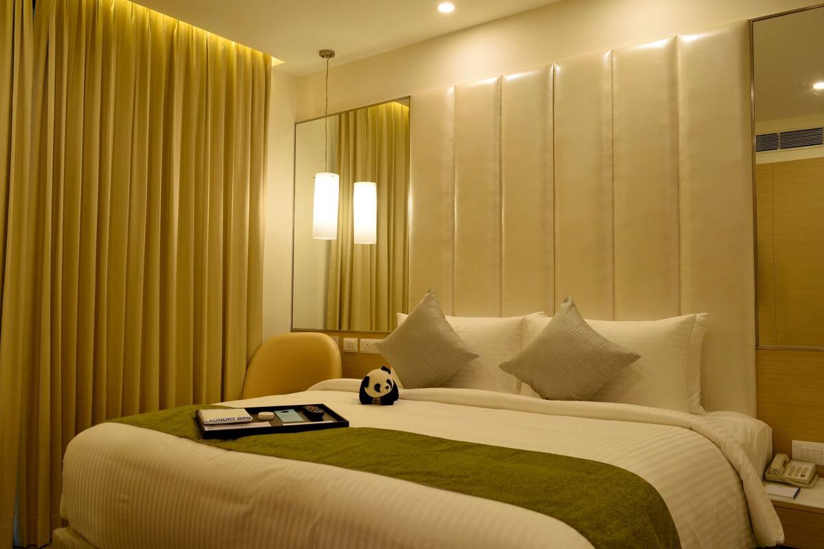 A hotel room with a neatly made bed, a green throw blanket, a panda plush toy, a remote control, and a phone on the nightstand.
