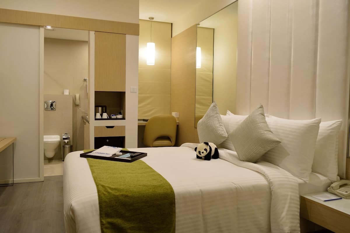A hotel room with a bed, a bedside table with a phone, a chair, a desk, and a mirror.
