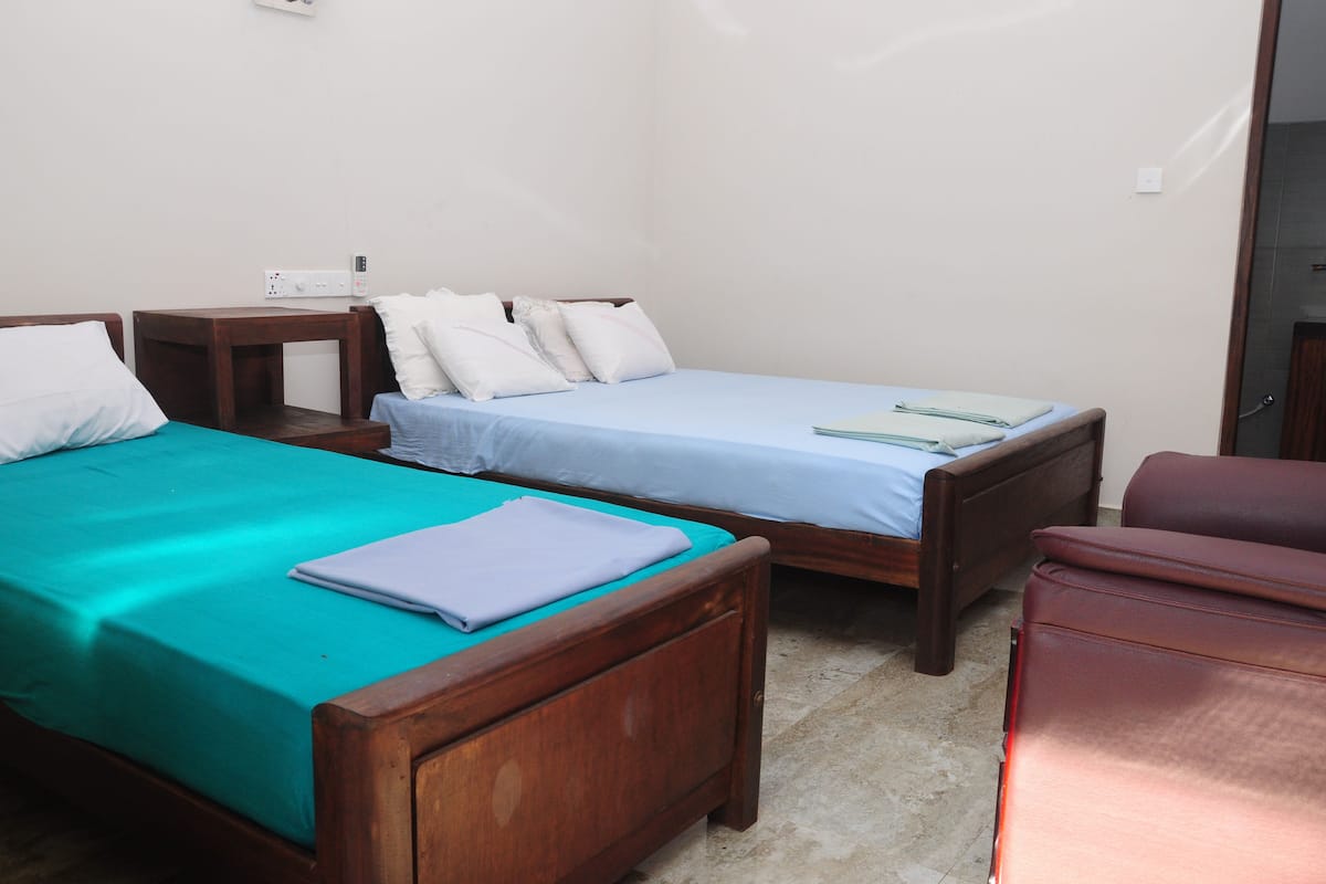 Una habitación con dos camas de madera, cada una con un edredón de color diferente. Hay una mesita de noche con un enchufe y una lámpara entre las camas. Se observa una silla roja en la esquina.