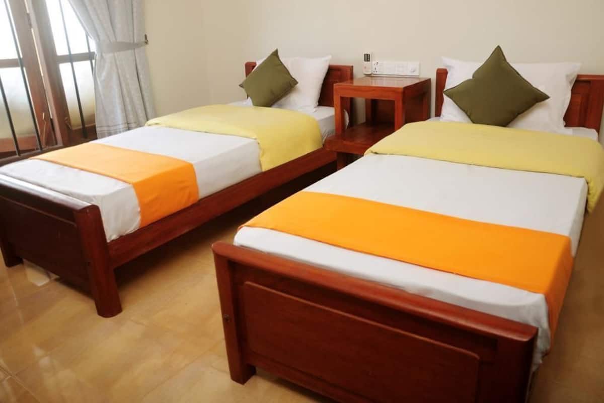 Dos camas de madera con ropa de cama blanca y naranja, una mesita de noche de madera, y una ventana con cortinas.