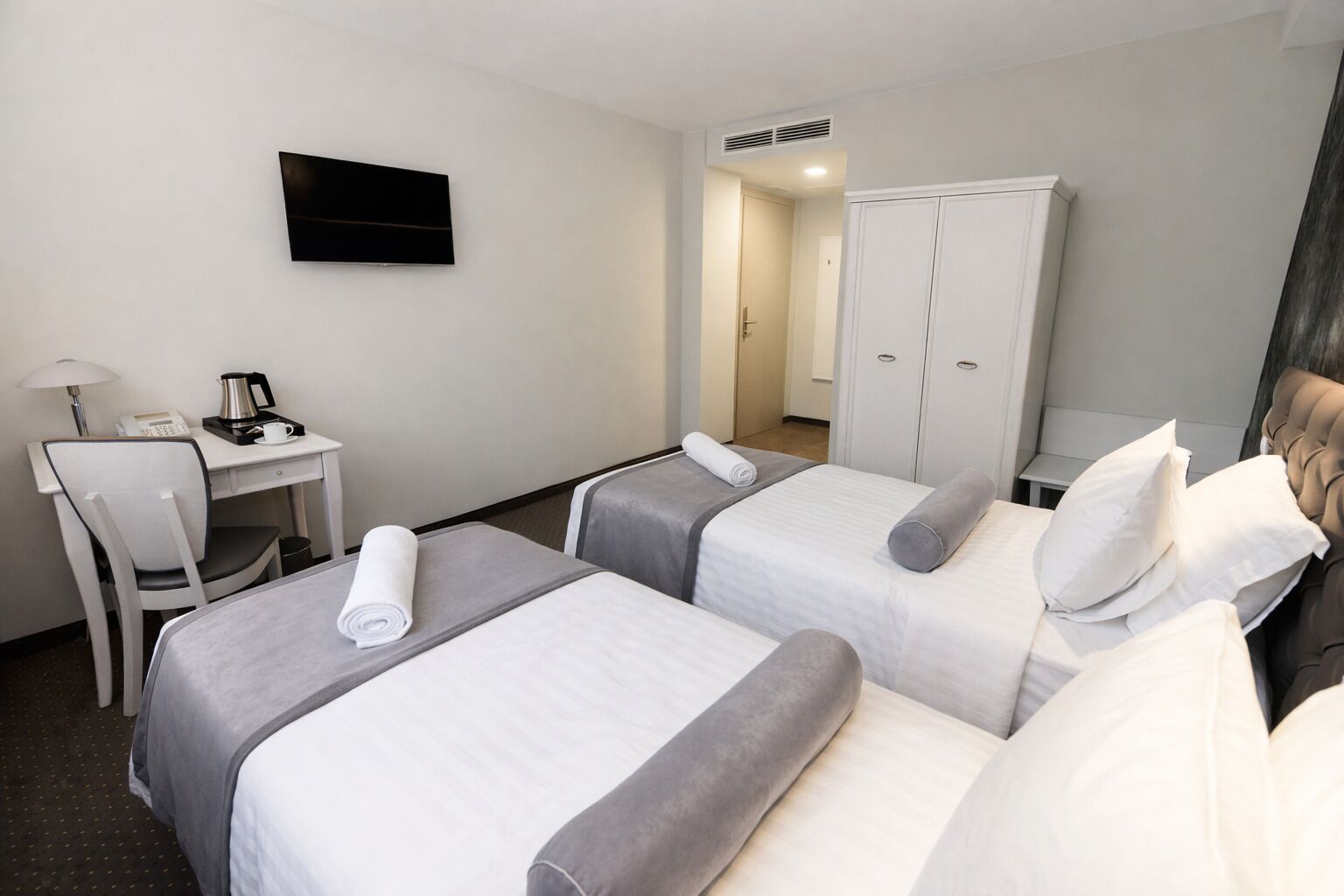 A hotel room with two beds, a desk with a telephone and coffee maker, a TV on the wall, and a wardrobe.