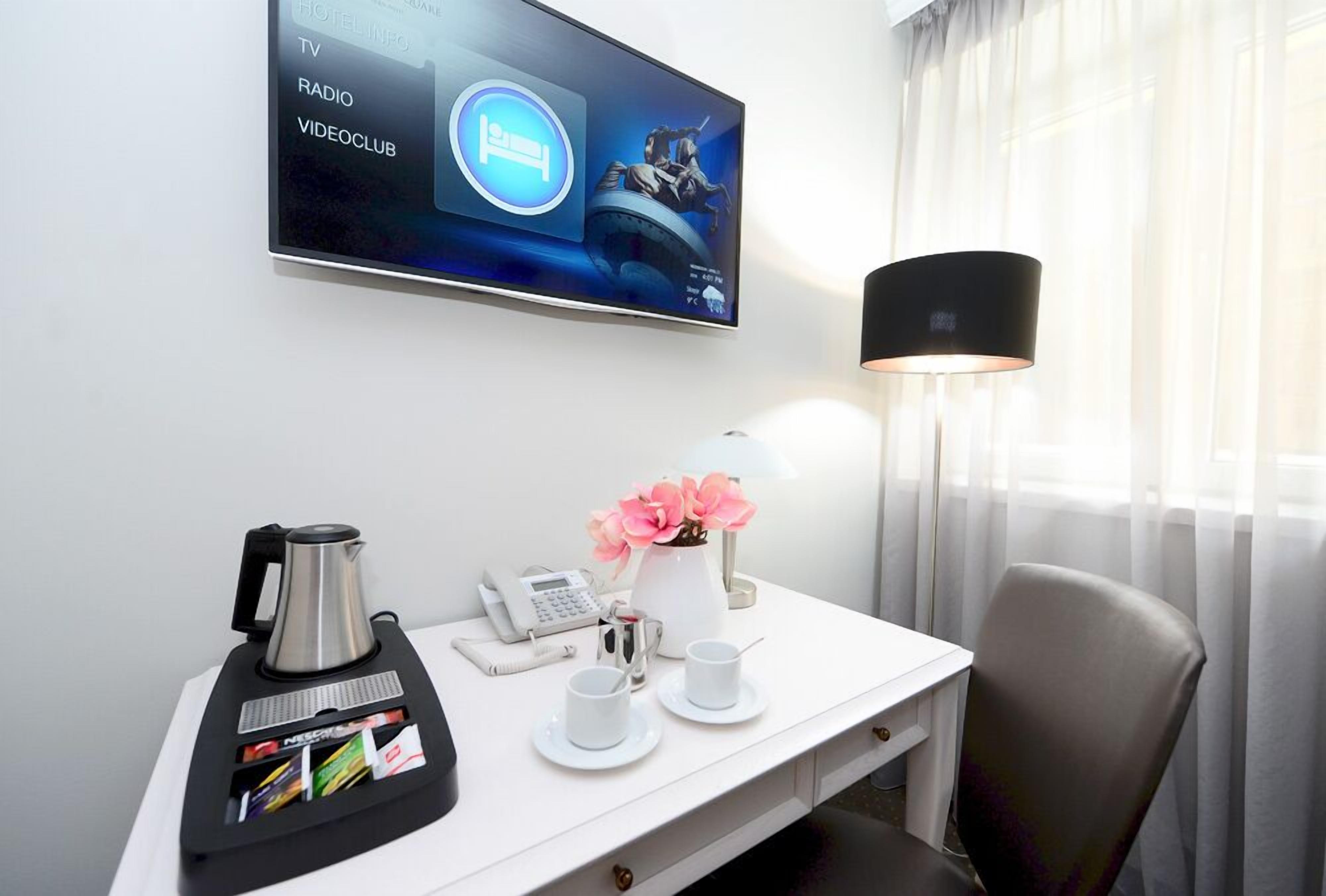 A hotel room with a flat-screen TV displaying hotel information, a coffee maker, a telephone, a vase with flowers, and a lamp.