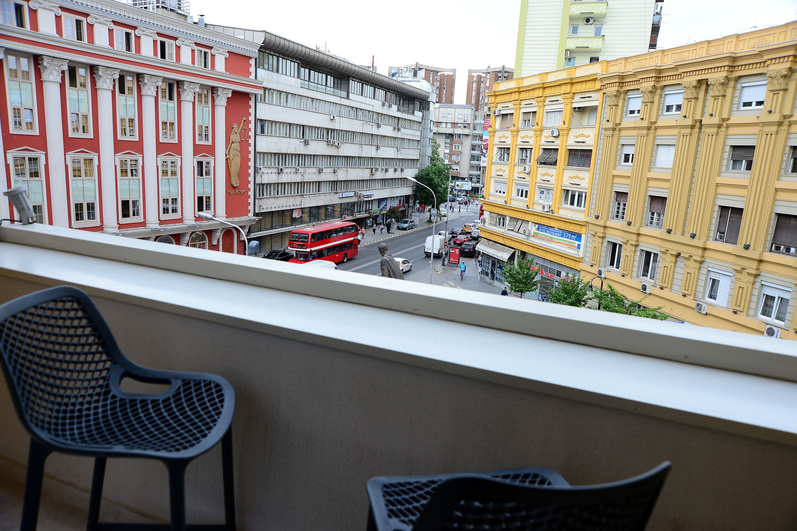 A balcony with a view of a city street, including a red double-decker bus and various buildings.