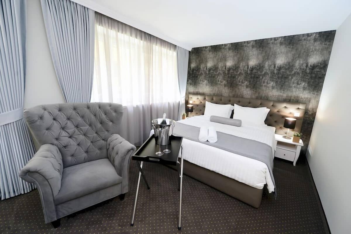 Une chambre d’hôtel moderne avec un canapé gris à motifs capitonnés, un lit soigneusement fait avec du linge de lit blanc, une table de chevet avec une lampe et une grande fenêtre ornée de rideaux légers.