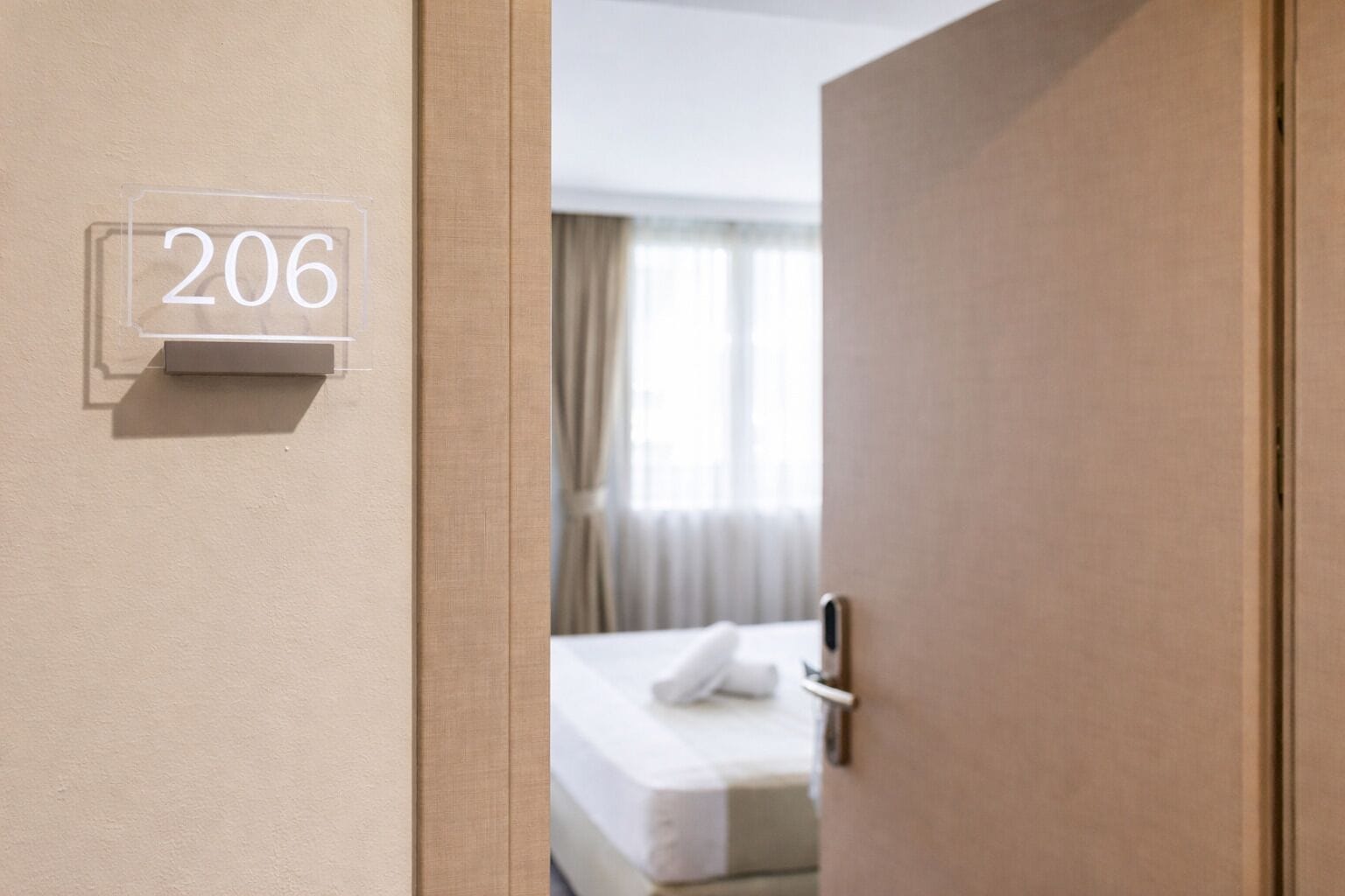 A hotel room with a door labeled '206,' a bed with a white mattress and a white pillow, and a window with curtains.