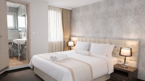 A hotel room with a bed, two bedside lamps, a nightstand, a mirror, and a bathroom visible through an open door.
