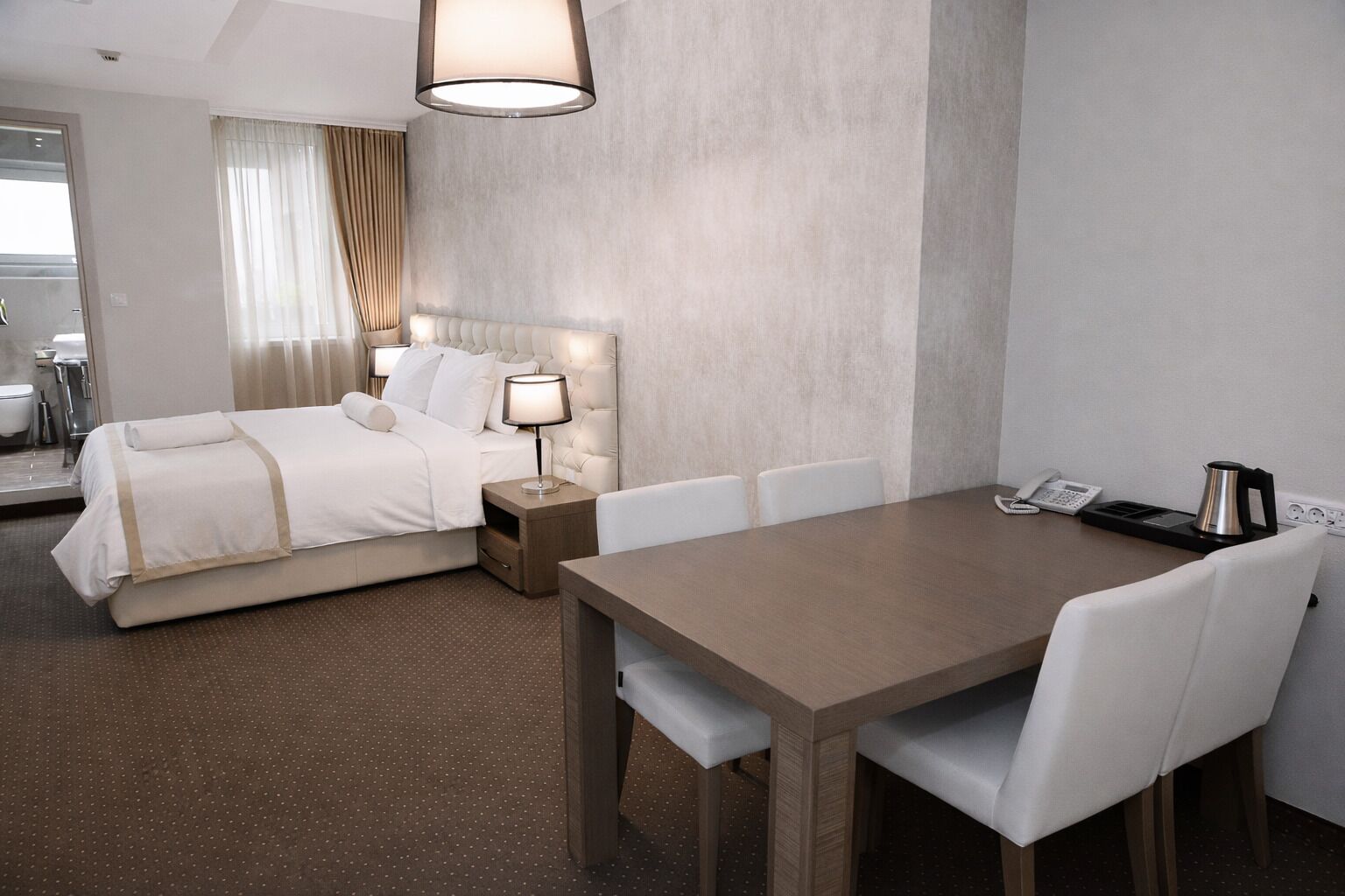 A hotel room with a bed, bedside table, lamp, and a dining table with chairs.
