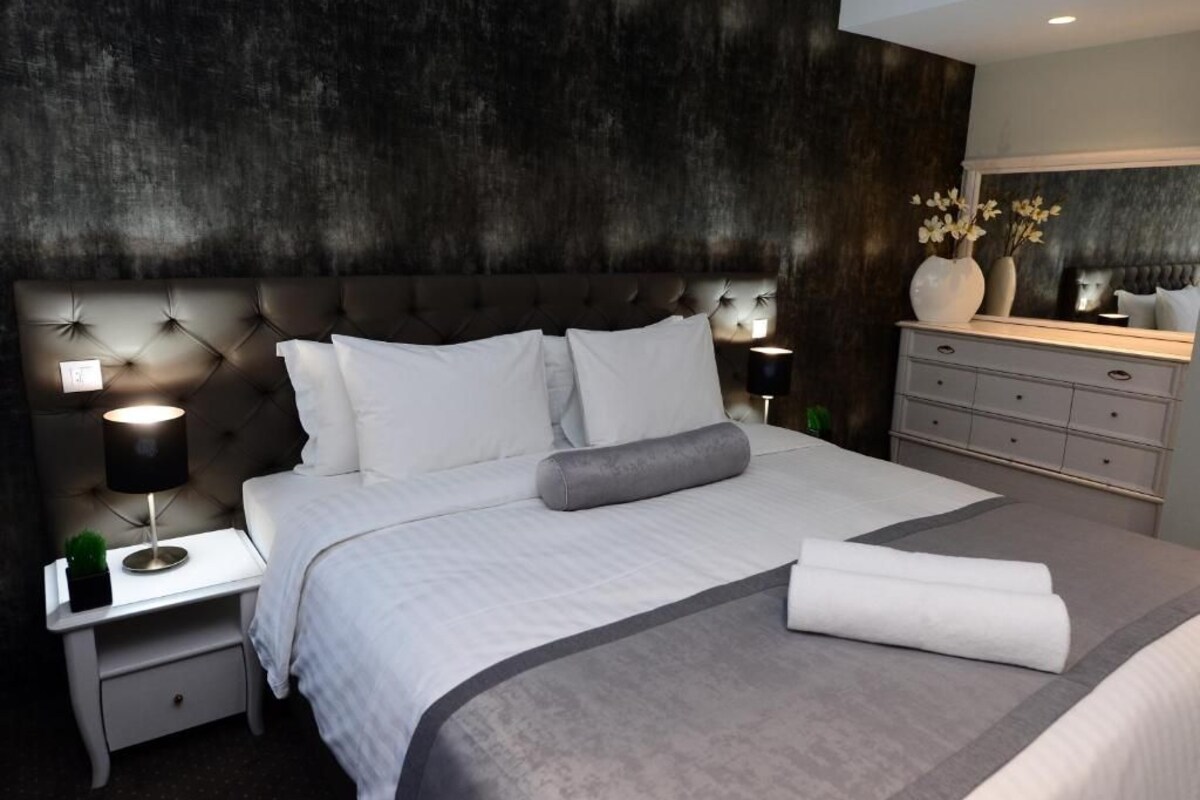 Un dormitorio moderno con cabecera capitonada, cama bien tendida con sábanas blancas, una lámpara de noche y un aparador con espejo.