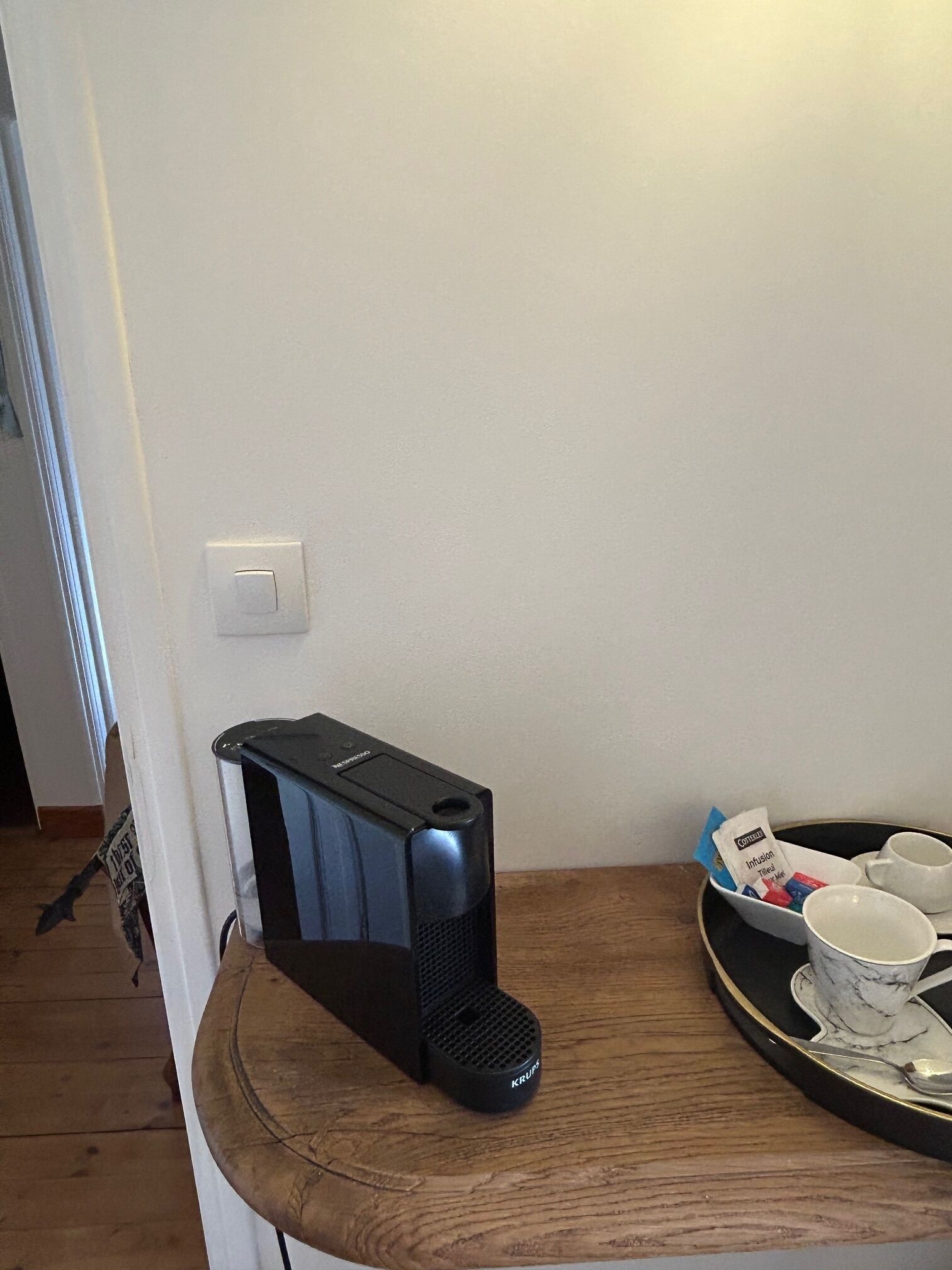 Una cafetera sobre una mesa de madera, con una bandeja de tazas y un paquete de cápsulas de café.