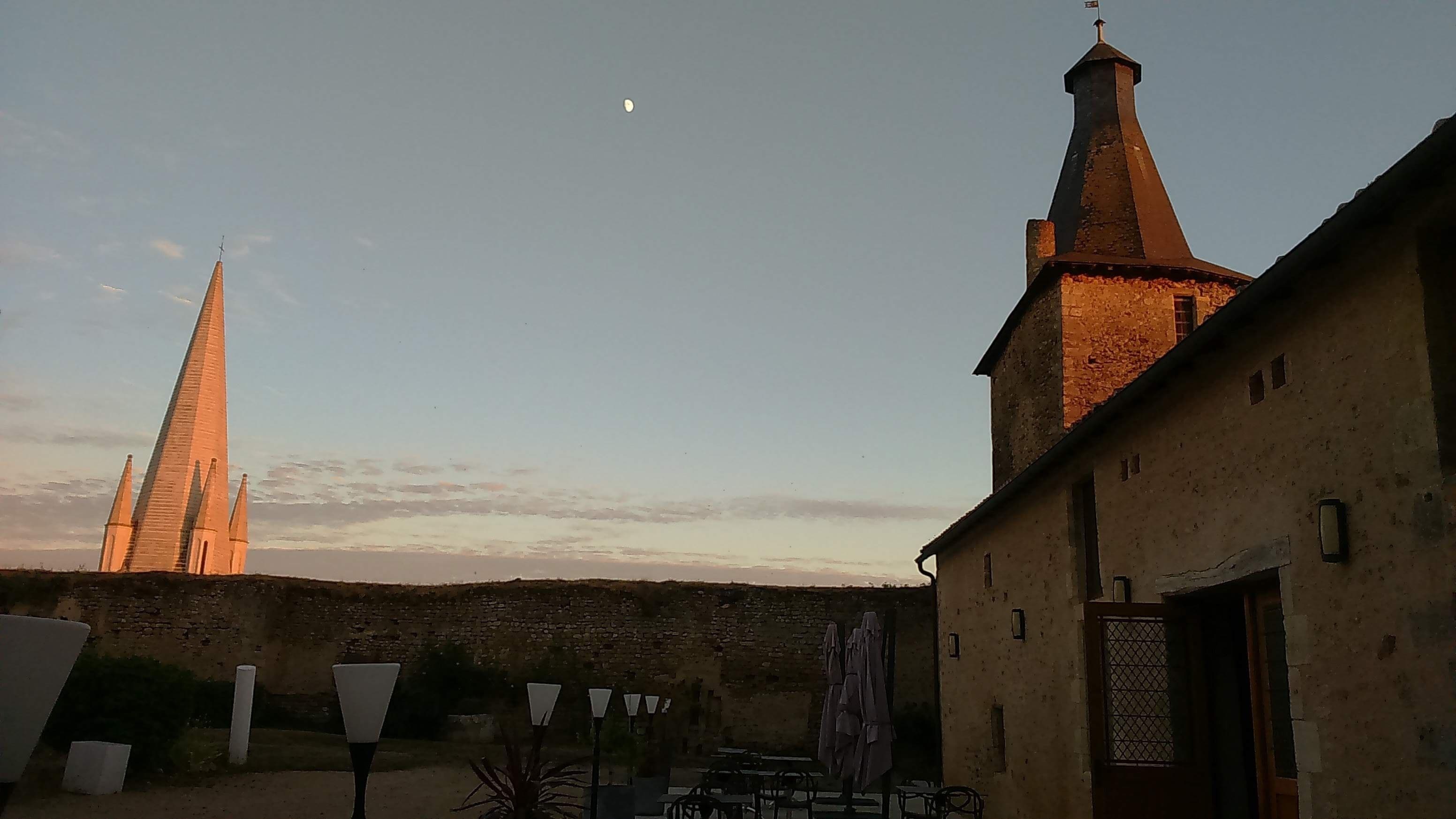 A building with a prominent tower and a smaller structure in the background. There are outdoor tables and chairs, and a lamp post is visible.