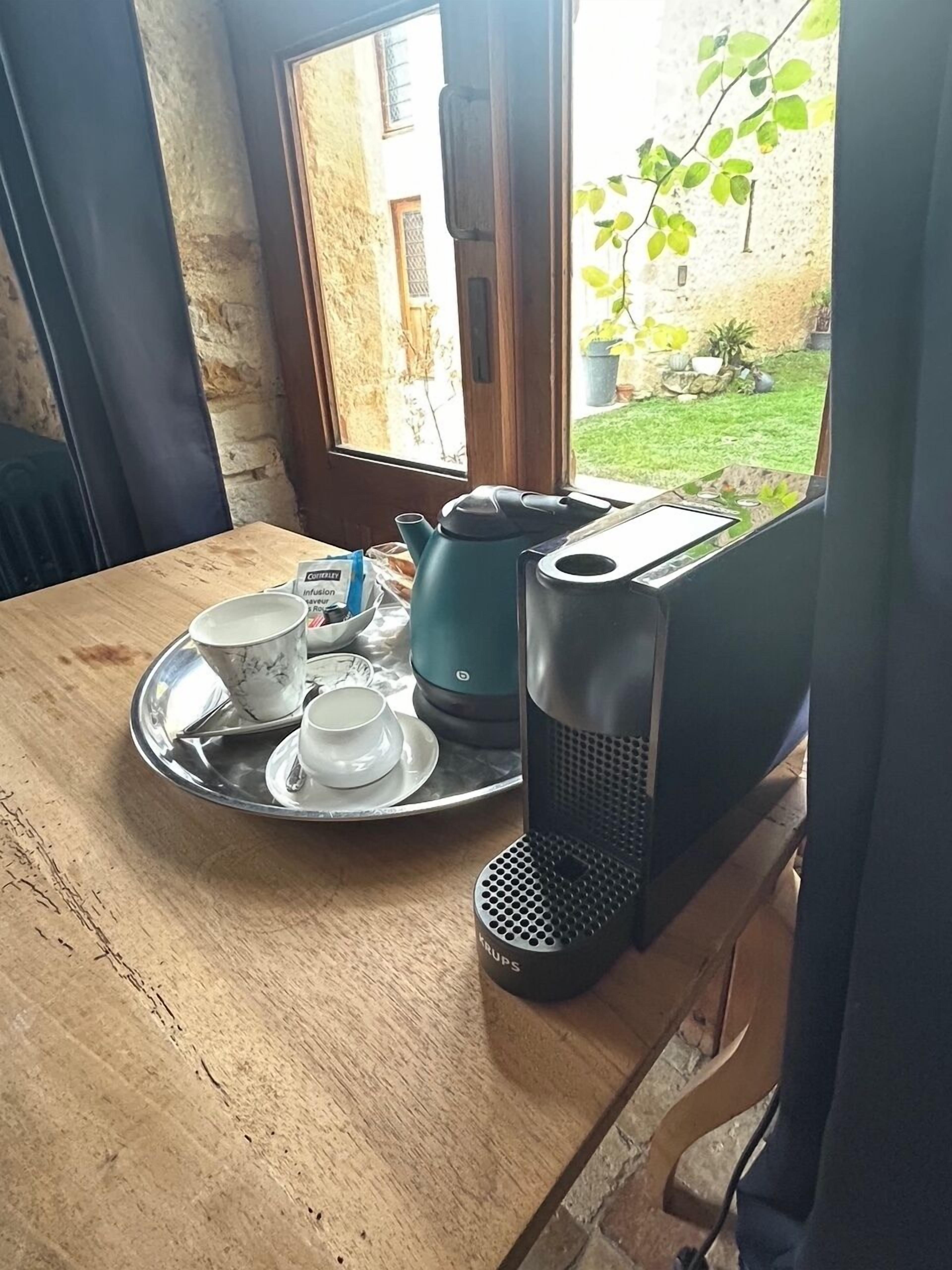 Un plateau de petit-déjeuner avec une théière, des tasses et une cafetière, posé sur une table en bois près d’une fenêtre.