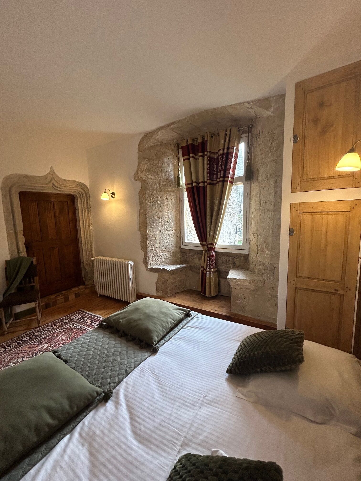 A bedroom with a bed, a chair, a radiator, a window with curtains, and wooden doors.