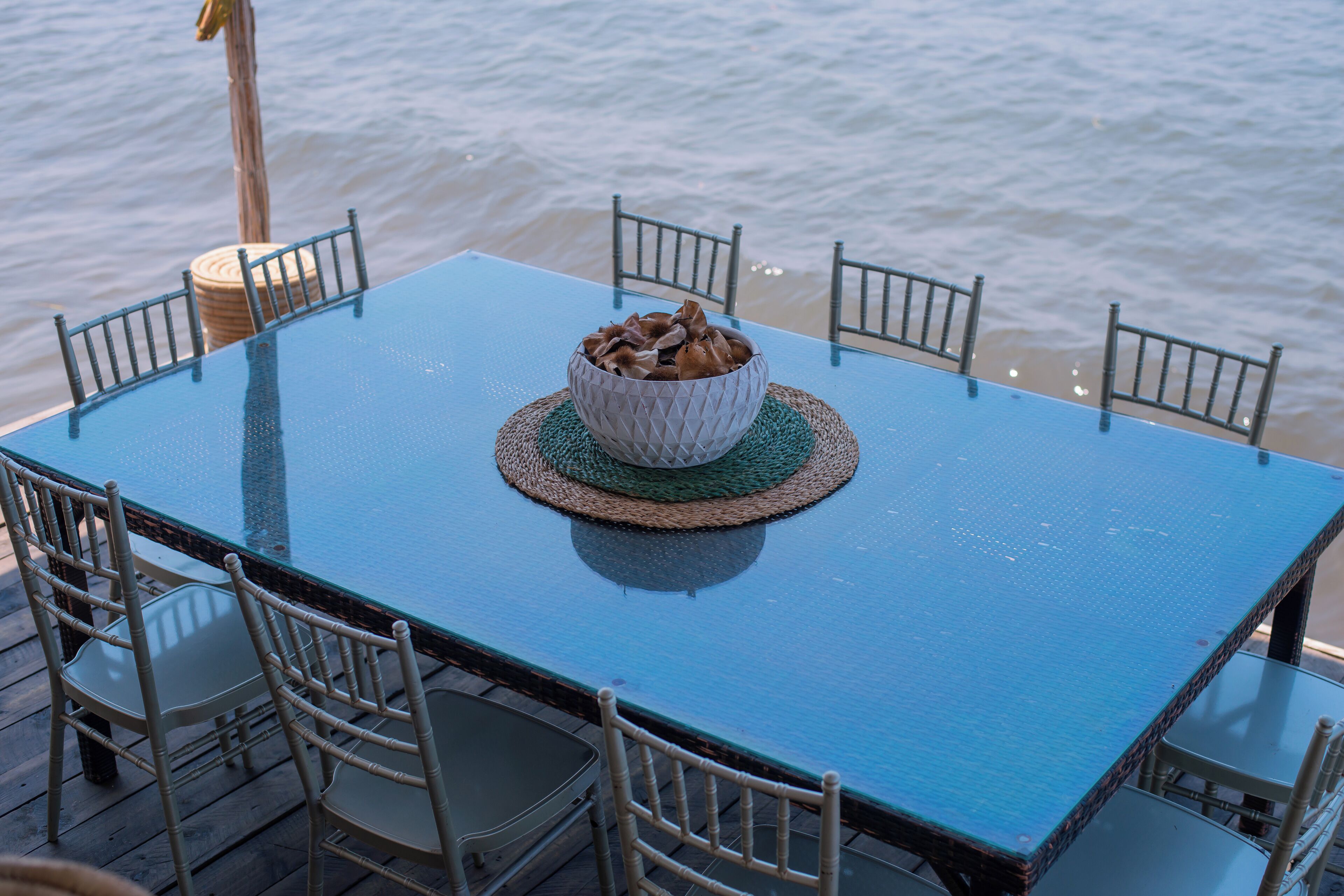 Une table avec un set de table en fibres tressées, un bol rempli de nourriture et six chaises, le tout placé sur une terrasse au bord de l’eau.
