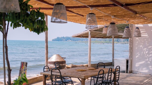 Area pranzo all'aperto con vista sul mare, caratterizzata da un tavolo e sedie in legno, lampade a sospensione e un tetto di paglia.