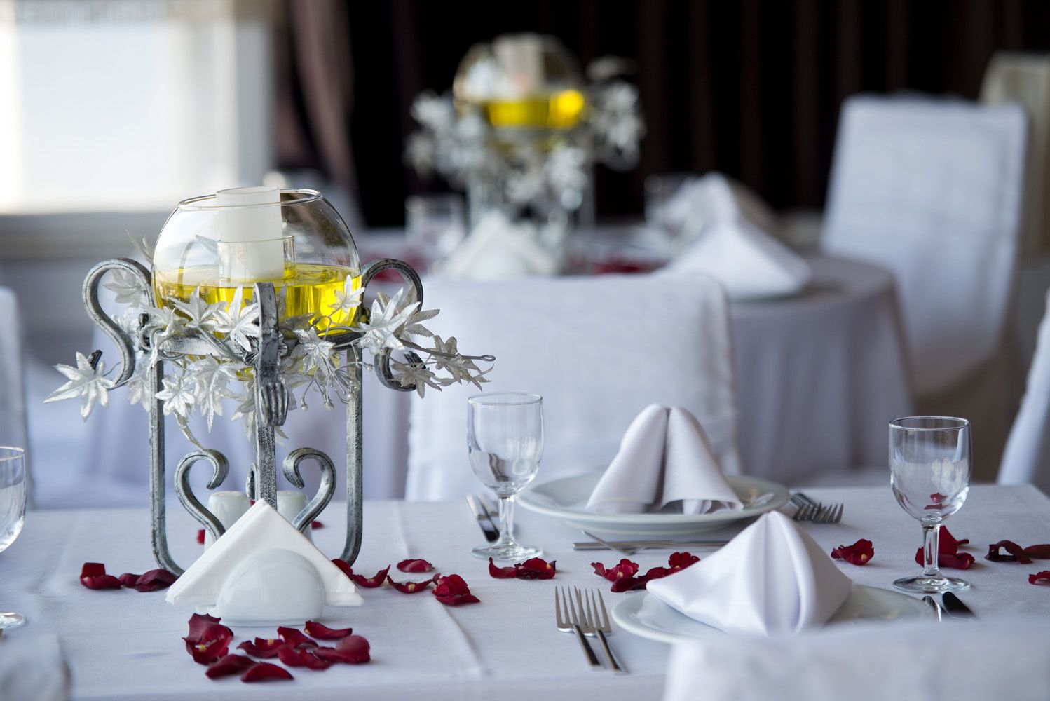 Mesa lista para comer con manteles blancos, cubiertos y un centro de mesa metálico decorado con flores.