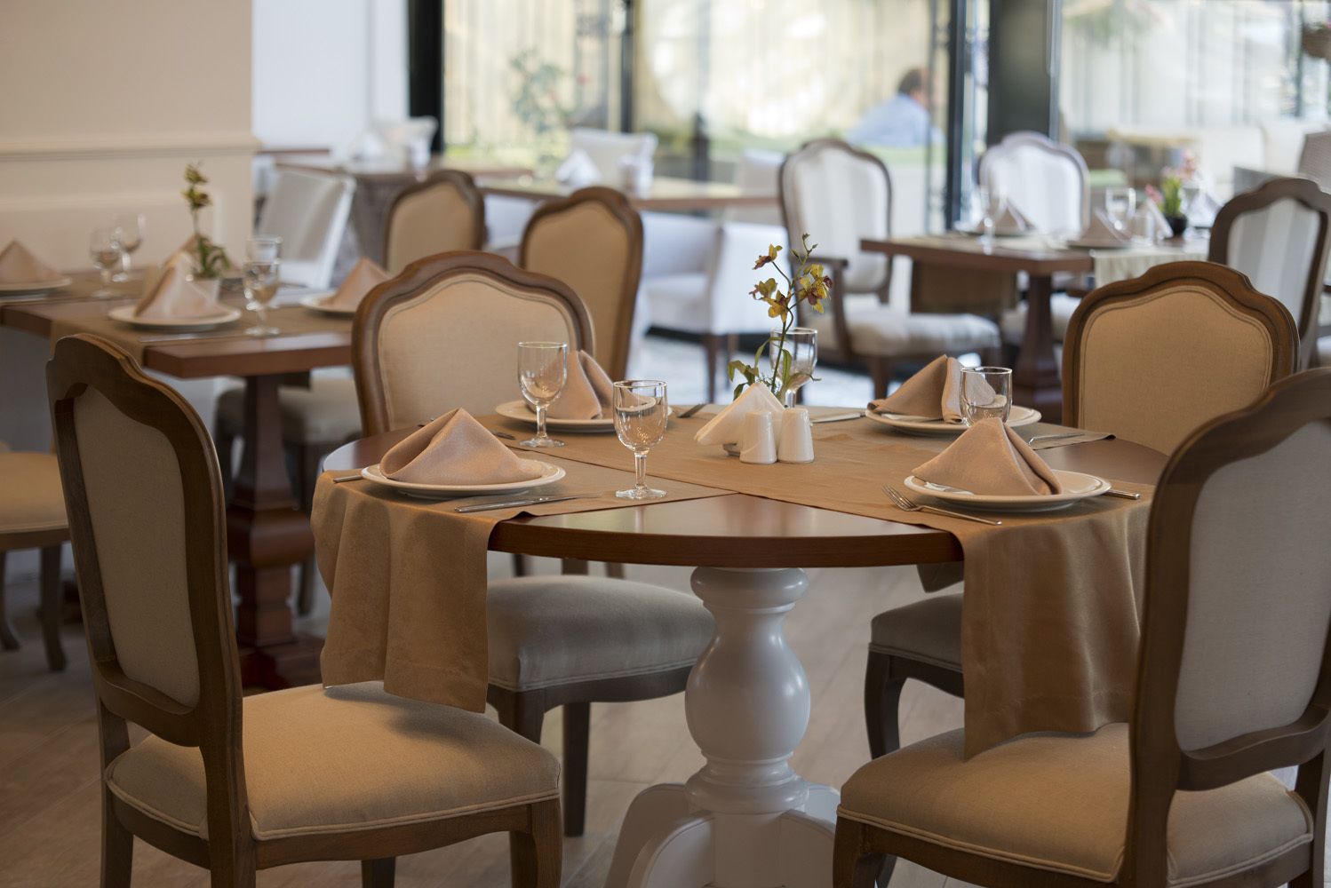 Un comedor con mesas redondas, sillas color beige y mesas cuidadosamente dispuestas con platos, vasos y servilletas.
