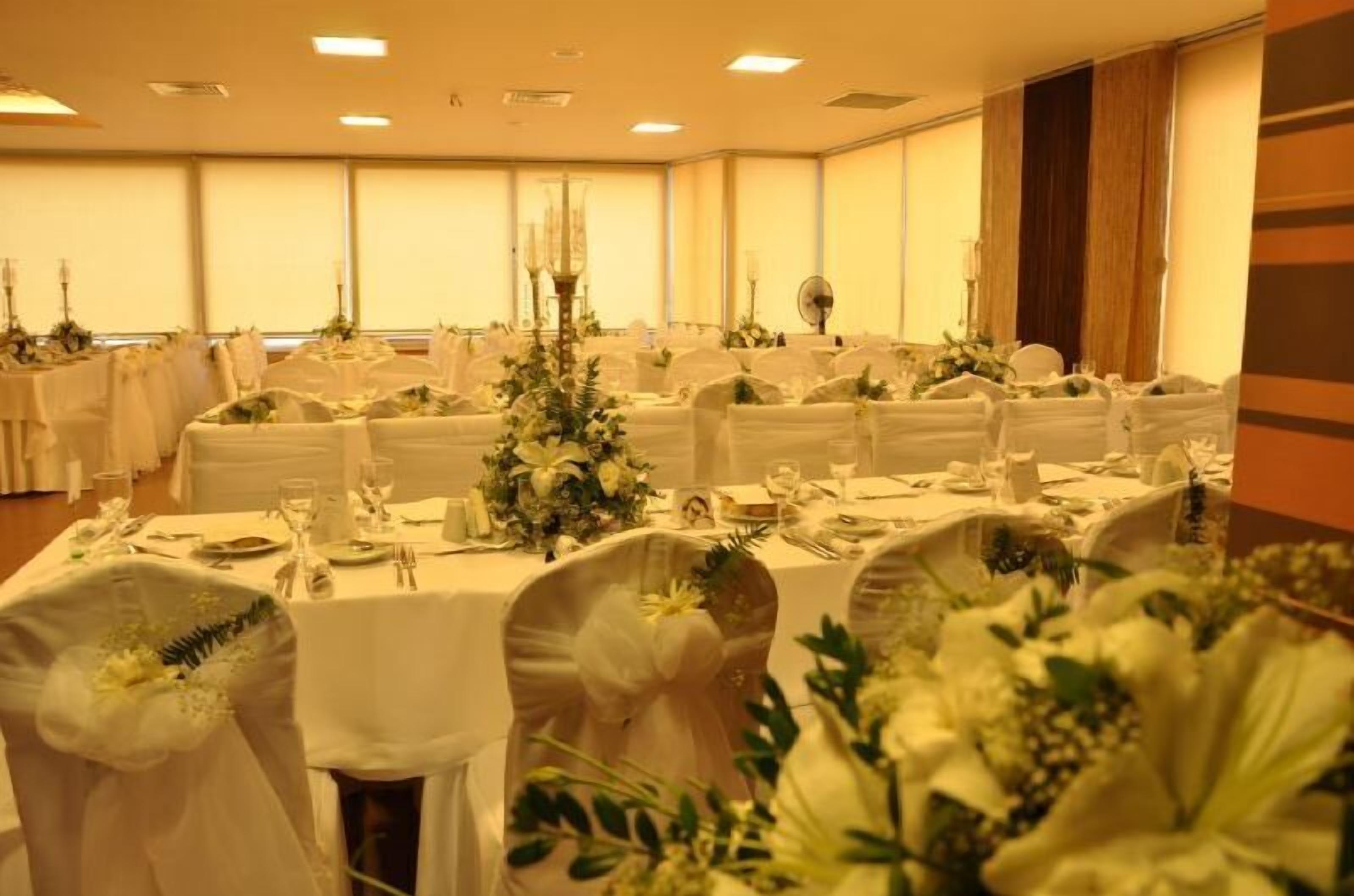 Un salón de banquetes con mesas redondas dispuestas para un evento formal, con manteles blancos, centros de mesa florales y sillas blancas adornadas con cintas.