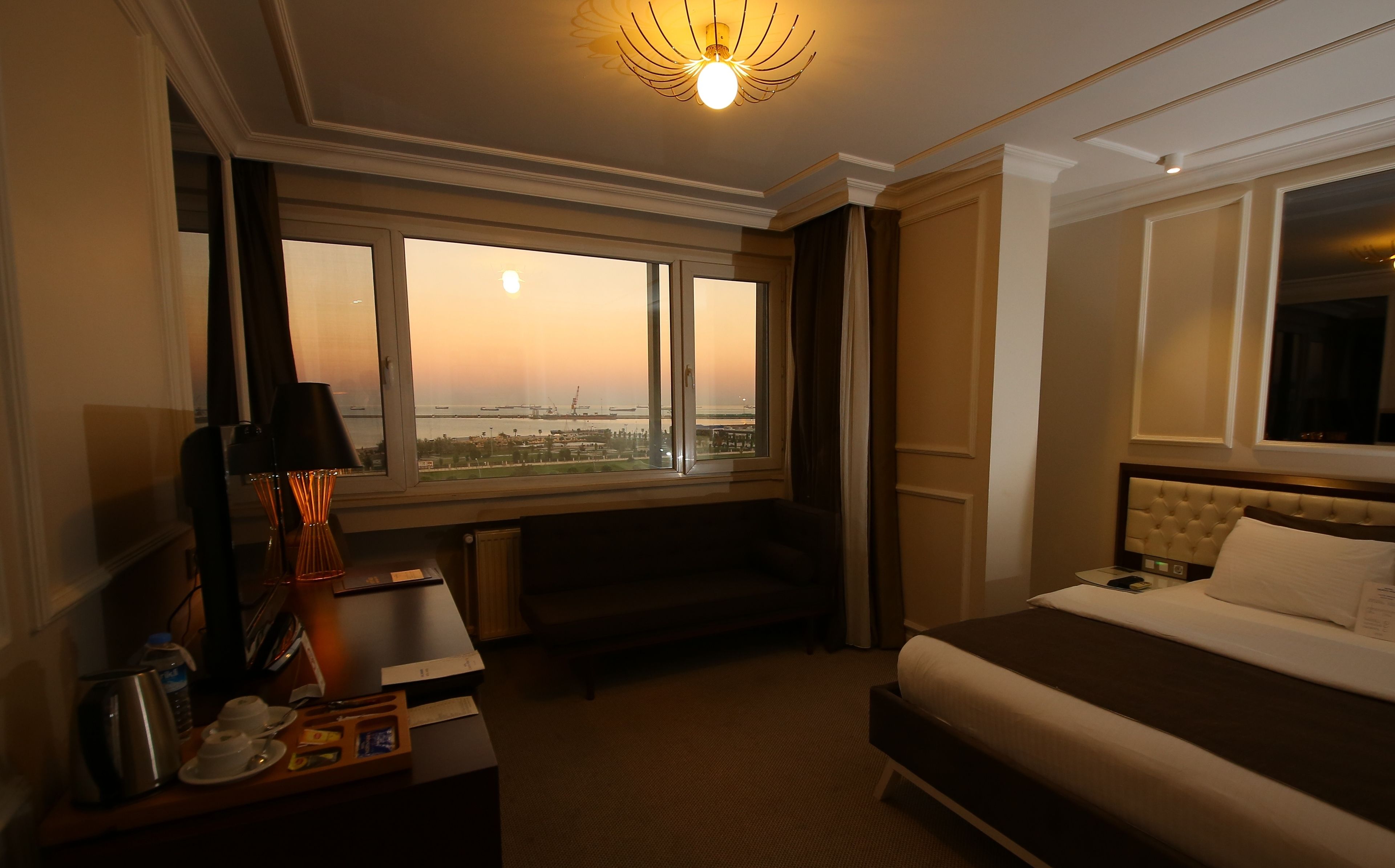 A hotel room with a large window offering a view of the cityscape, a bed, a bedside table with a lamp, and a small seating area.