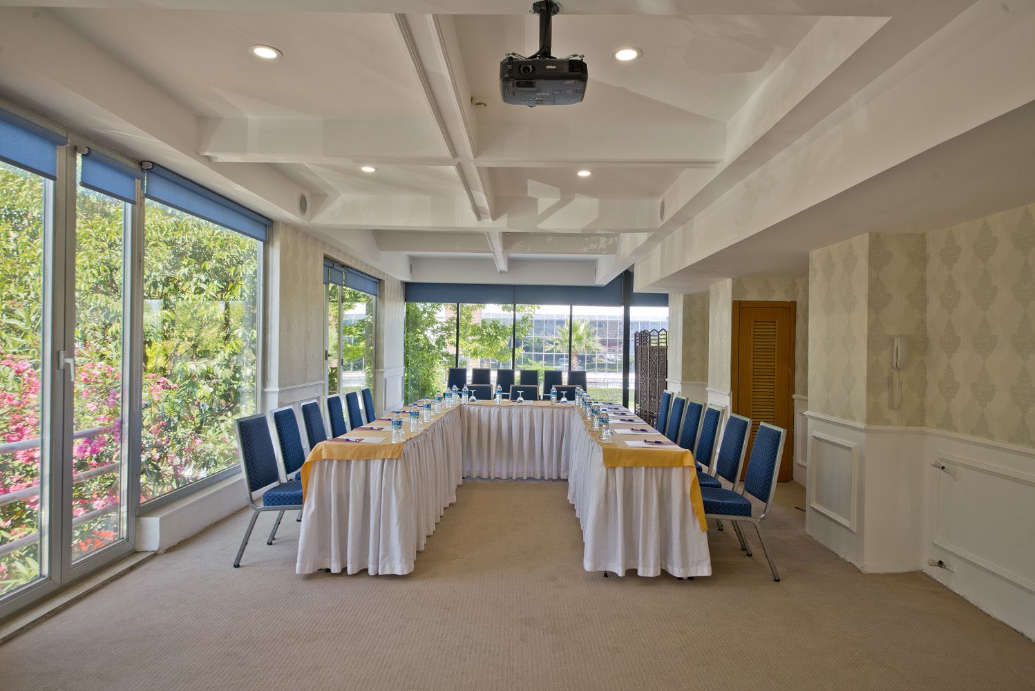 A conference room with a long table, blue chairs, and large windows offering a view of greenery.