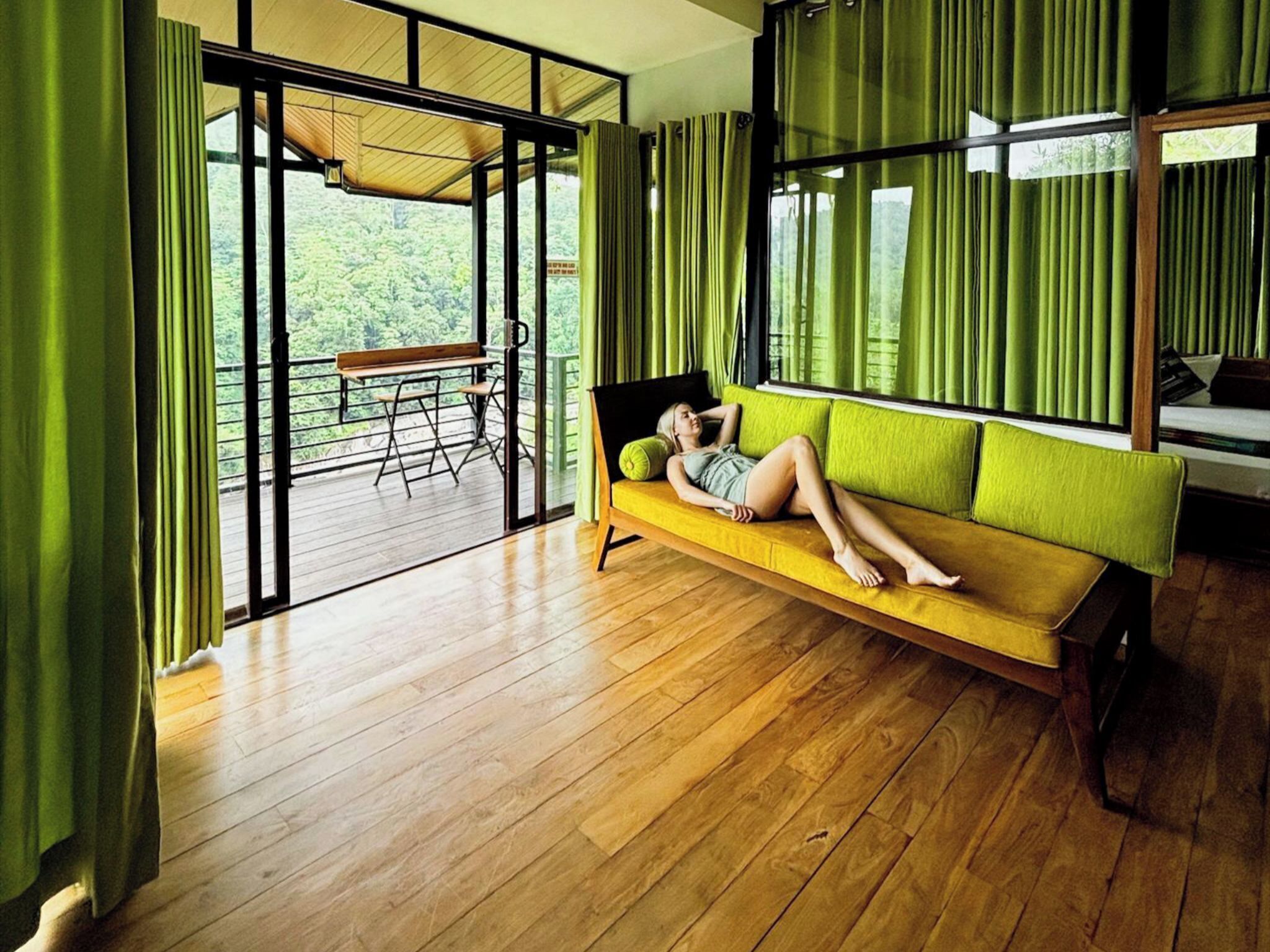 A room with a wooden floor, a yellow sofa, and a view of greenery through large windows.
