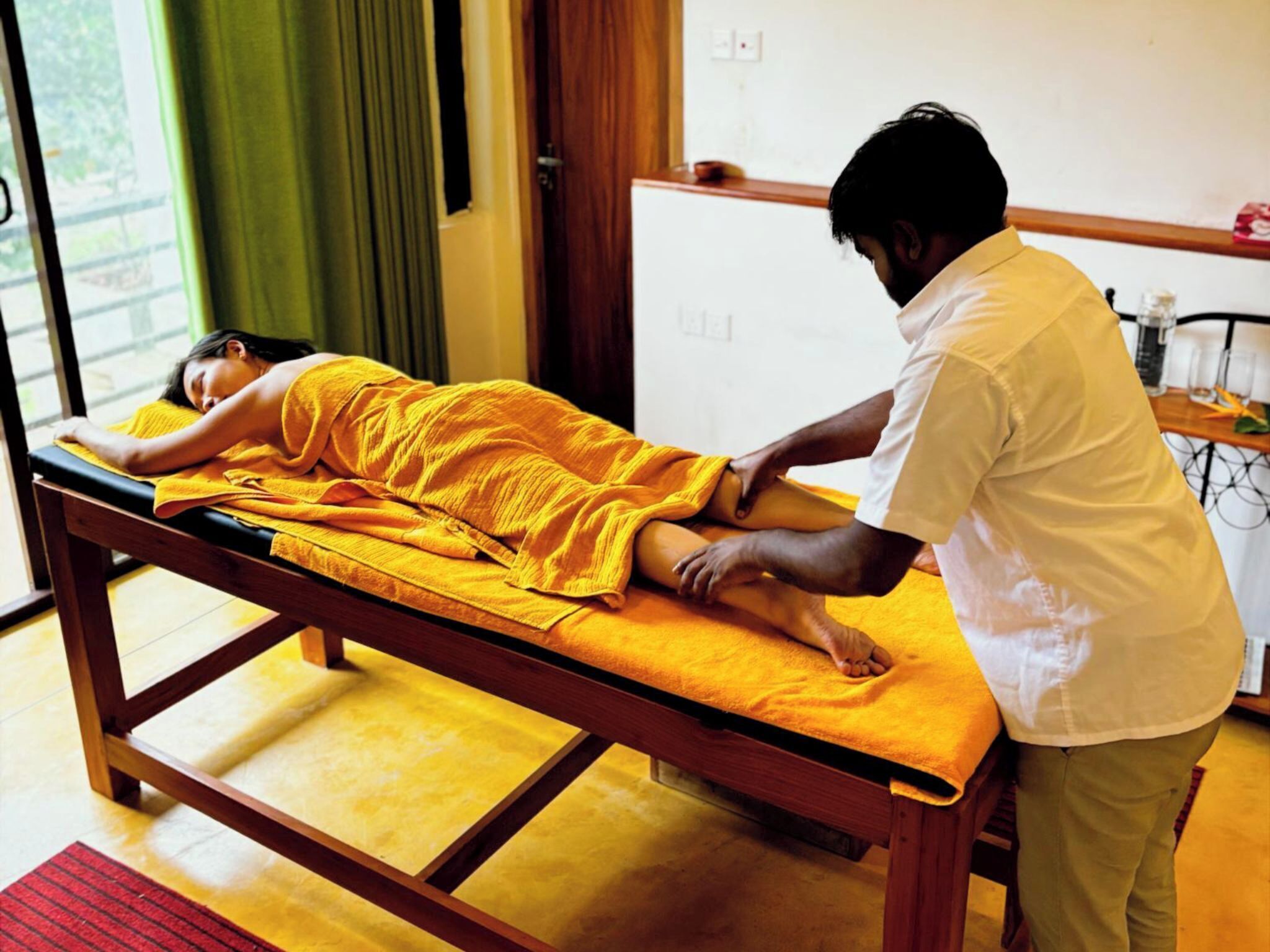 A massage therapy room with a wooden table, a person receiving a massage, and a small table with bottles in the background.
