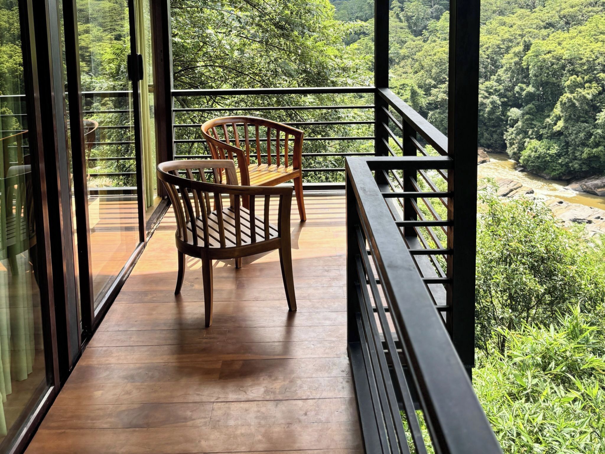 A balcony with wooden flooring and two wooden chairs, overlooking a lush green landscape with a river.