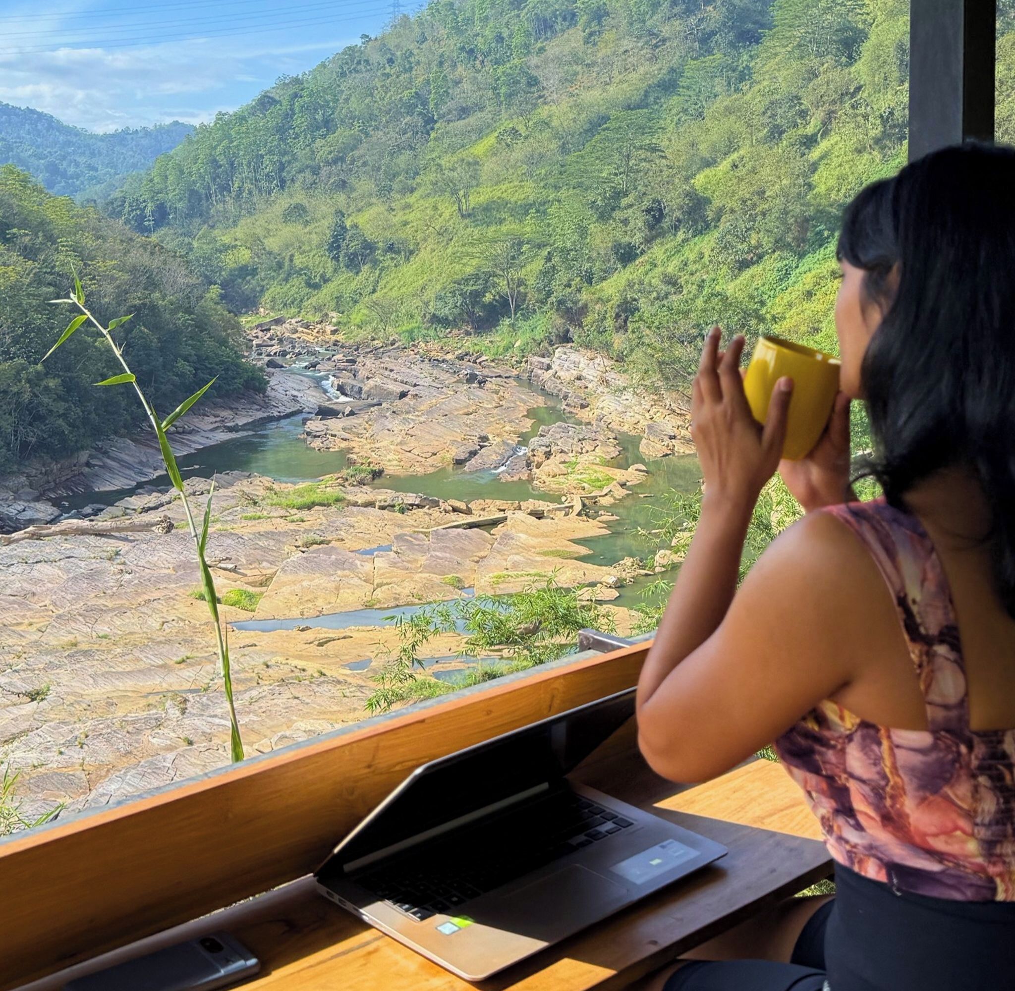 A person sitting by a window with a laptop, overlooking a river and forested hills.