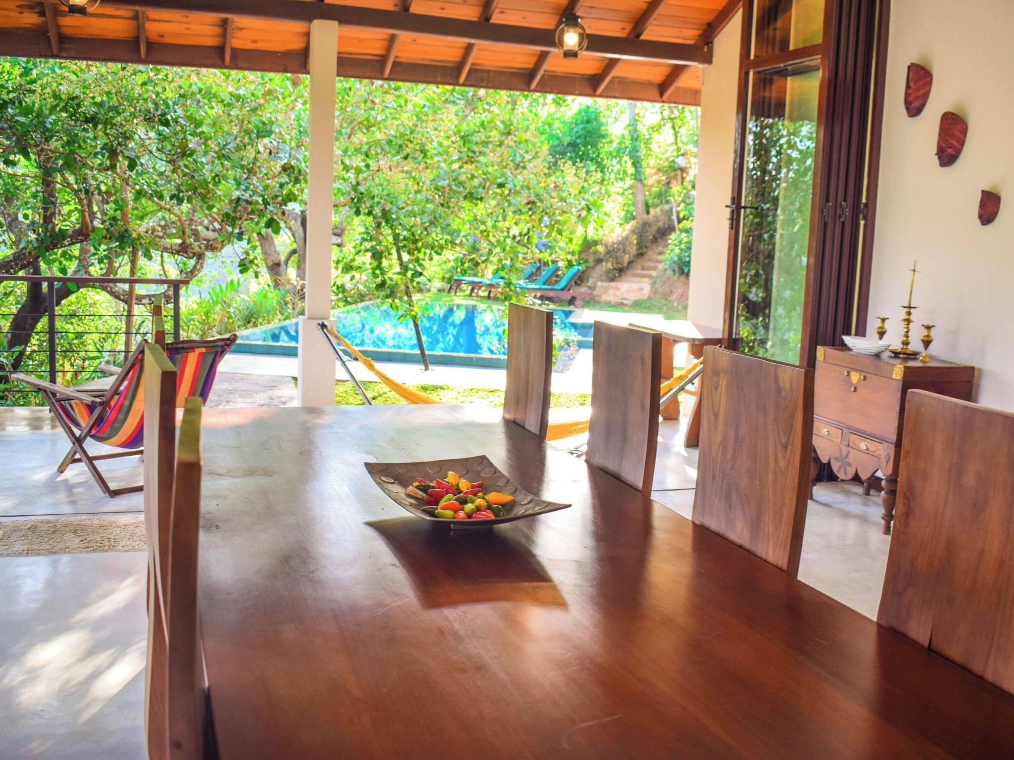 A wooden table with a fruit bowl, a hammock, and a view of a swimming pool and greenery.