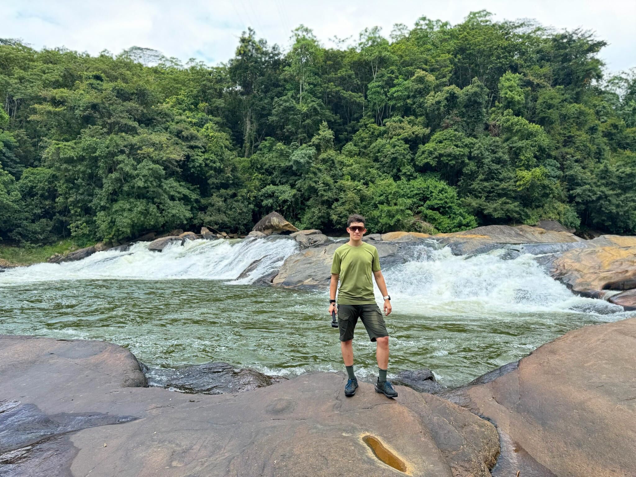 A person standing on a rock by a waterfall with dense forest in the background.