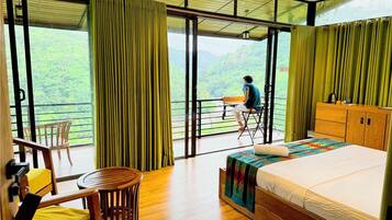 Una habitación con cama, una mesa, una silla y vistas a un paisaje verde y exuberante a través de amplios ventanales.