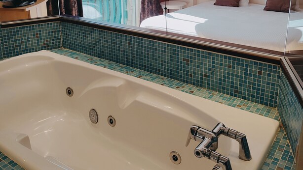 A bathroom with a large bathtub, a mirror, and a view of a bedroom with a bed and pillows.
