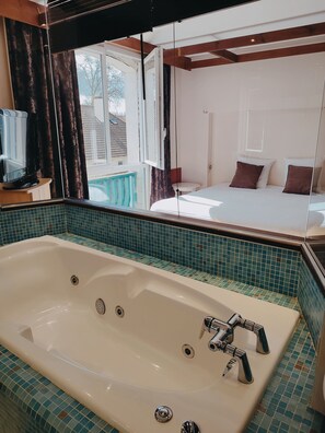 A bathroom with a large bathtub, a mirror, and a view of a bedroom with a bed and pillows. - Logis Hôtel Les Pages (Luneville)