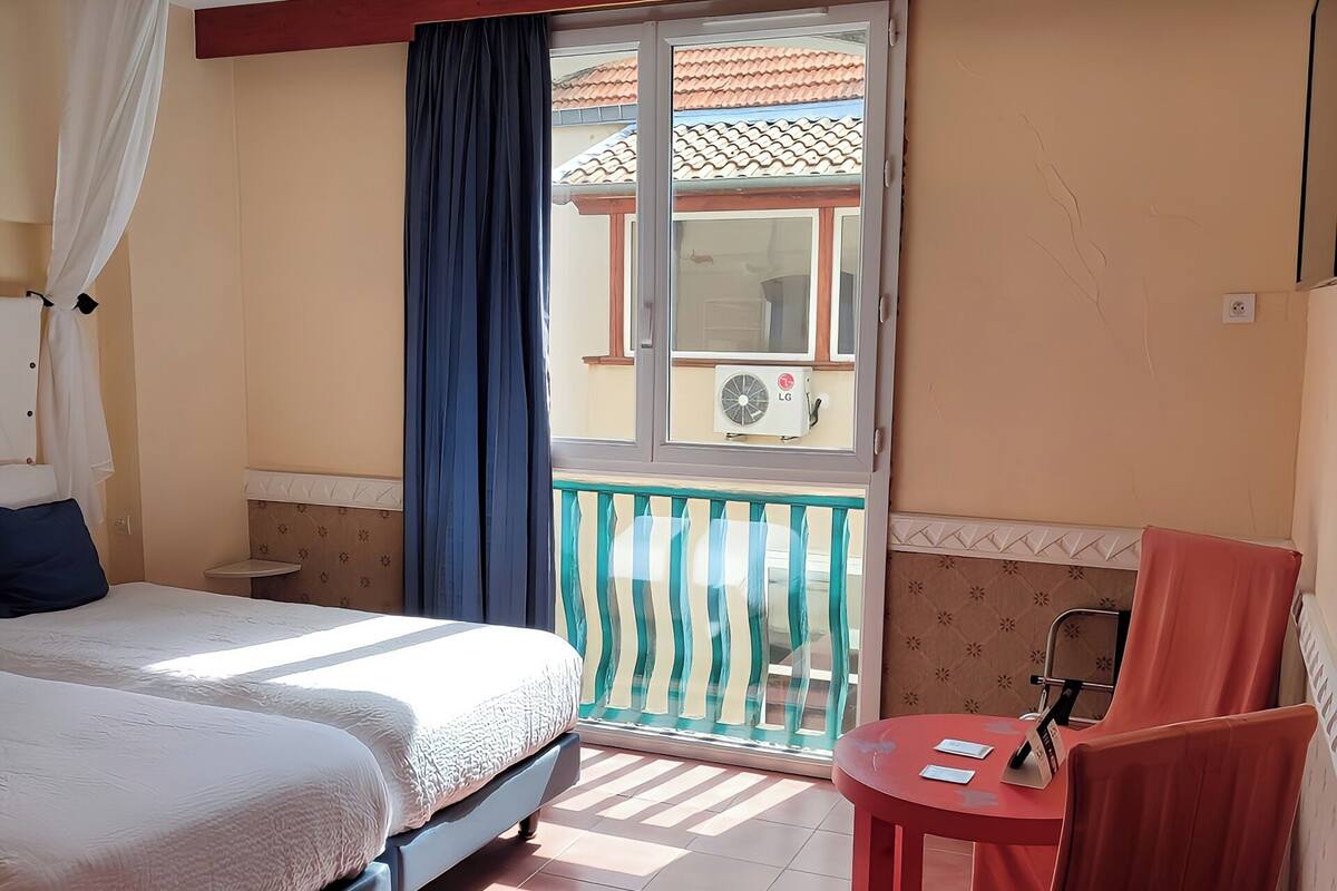A hotel room with a bed, a small table, a chair, a balcony with a view, and an air conditioning unit.