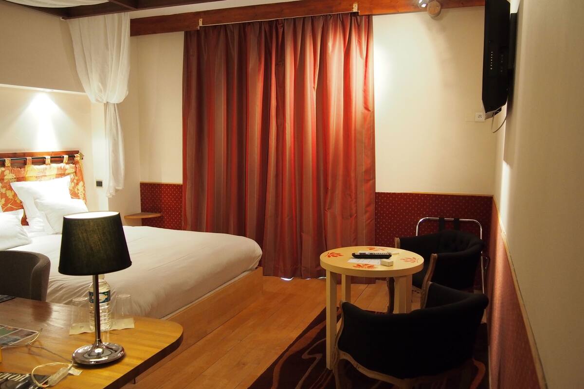 A hotel room with a bed, a desk with a lamp, a chair, a small table, and a TV mounted on the wall.