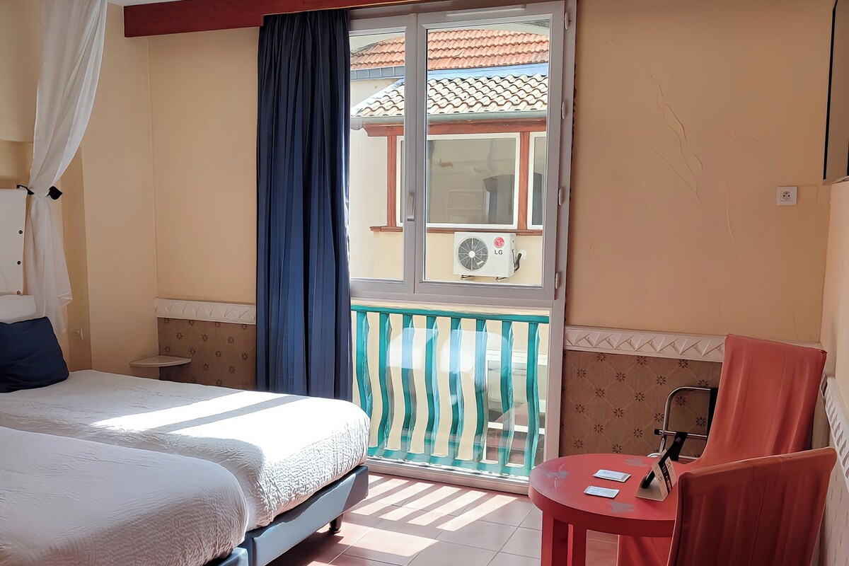 A hotel room with a bed, a small table, a chair, a balcony with a view, and an air conditioning unit.