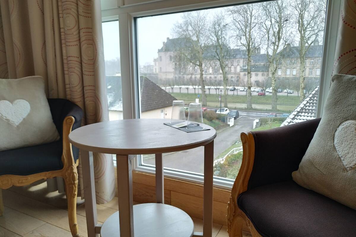 A room with a round wooden table, two upholstered chairs with heart-shaped pillows, a large window with a view of buildings and trees, and a curtain with a circular pattern.