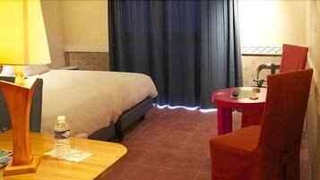 Une chambre d’hôtel avec un lit, un bureau avec une lampe, une chaise, une petite table avec une télécommande, et une salle de bain visible à travers une fenêtre.