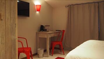 A hotel room with a bed, a desk, two red chairs, a television mounted on the wall, and a red wall light.